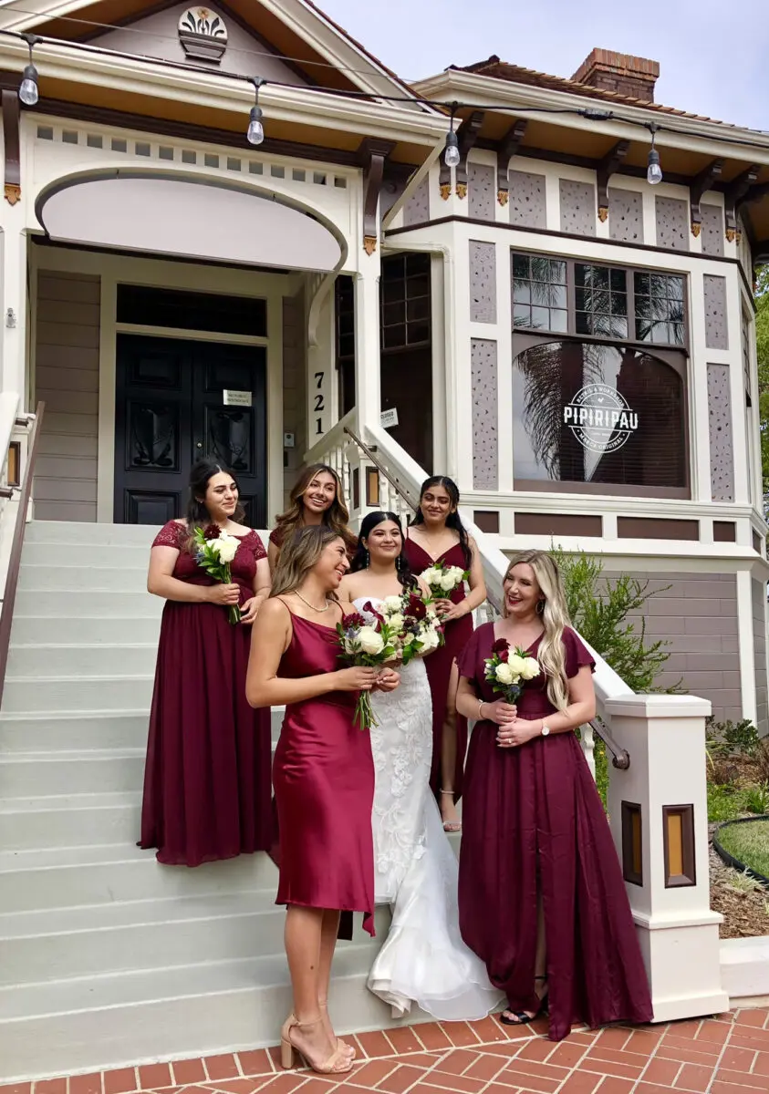 Bride in white gown with four bridesmaids in maroon dresses, posing on steps outside a vintage house in Heritage Square, Oxnard.