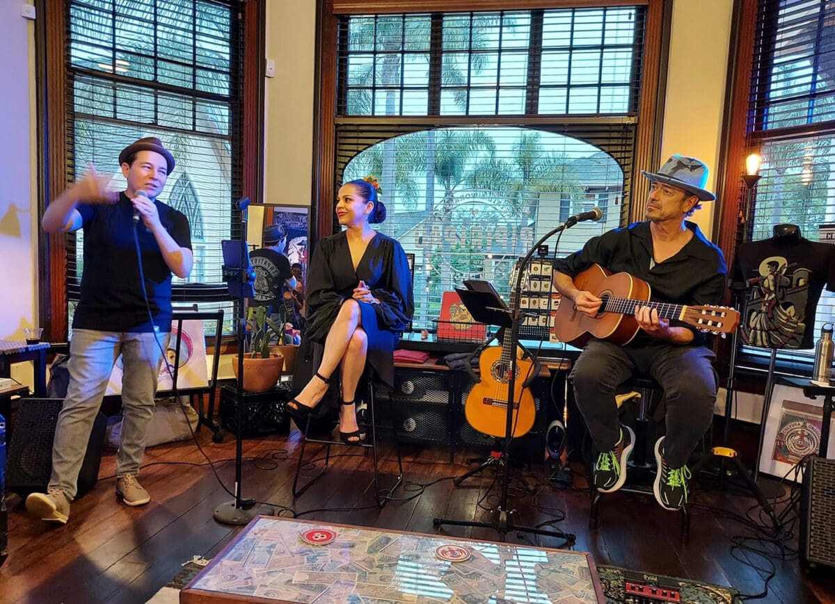 Three musicians performing in a cozy room with large arched windows at Heritage Square, including a singer, a guitarist, and a woman seated with a microphone.