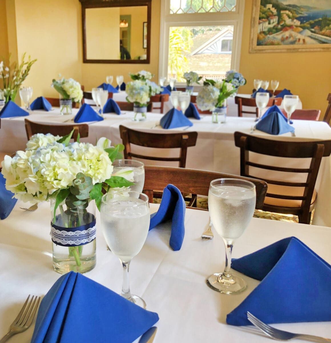 Elegant dining setup with blue napkins, white tablecloths, floral centerpieces, and glasses of water, in a room with bright windows and paintings at Heritage Square.