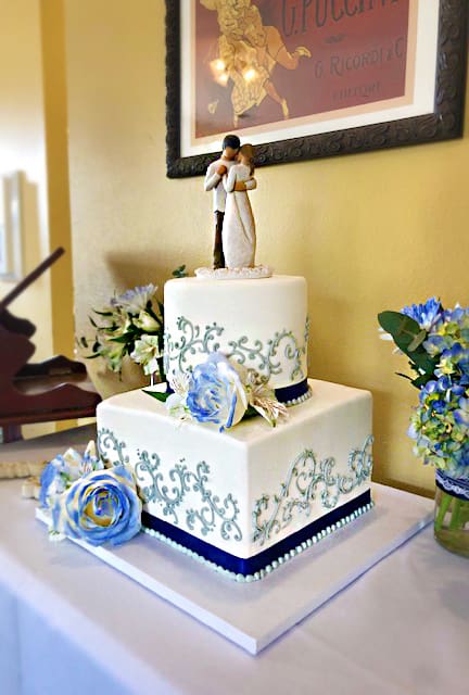 A wedding cake with a black couple figurine on top, decorated with blue flowers and elegant green patterns, displayed on a table at Heritage Square.
