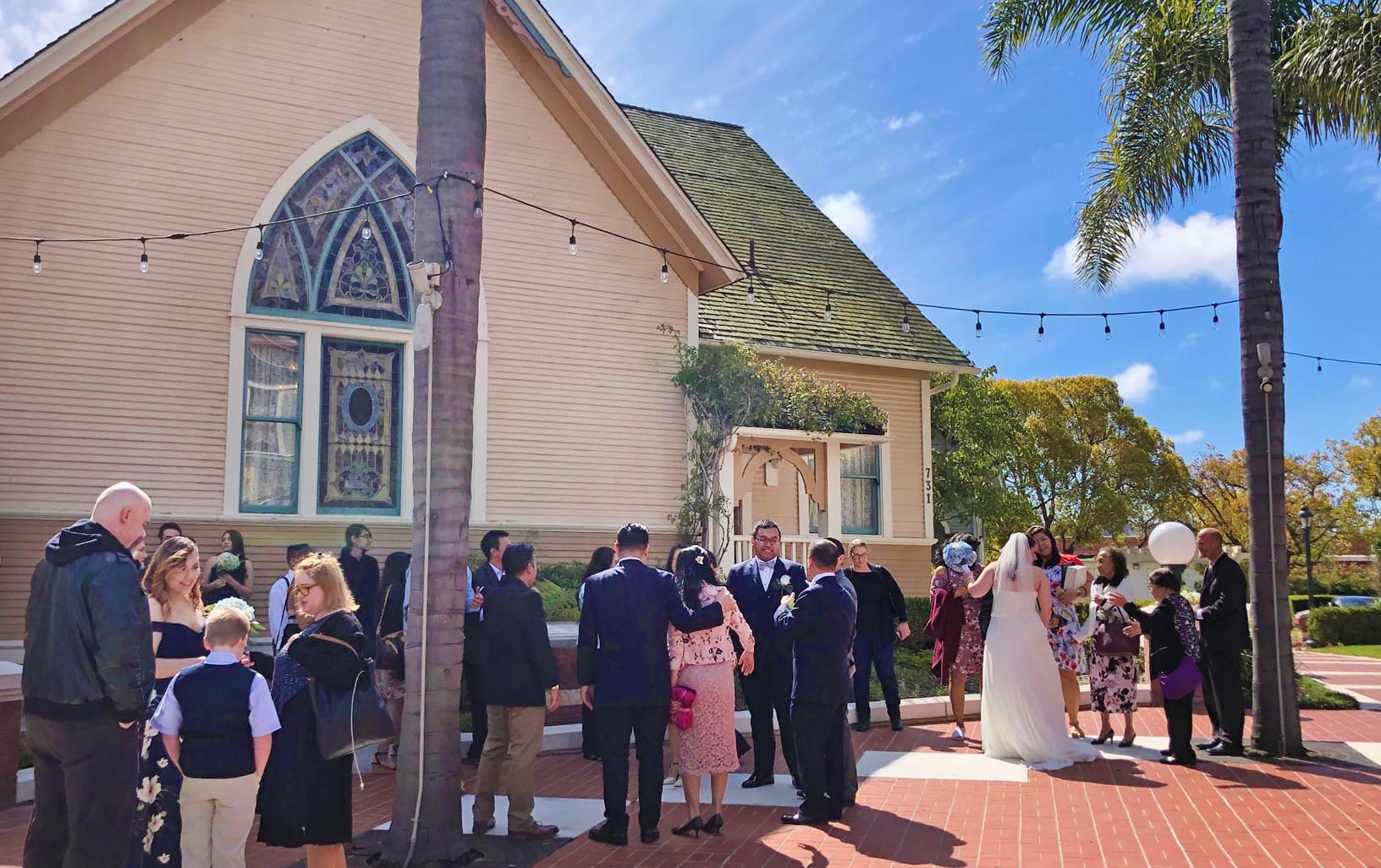 Wedding guests gather outside a quaint chapel in Heritage Square, Oxnard, under a clear sky, with a bride and groom in the center, surrounded by palm trees.