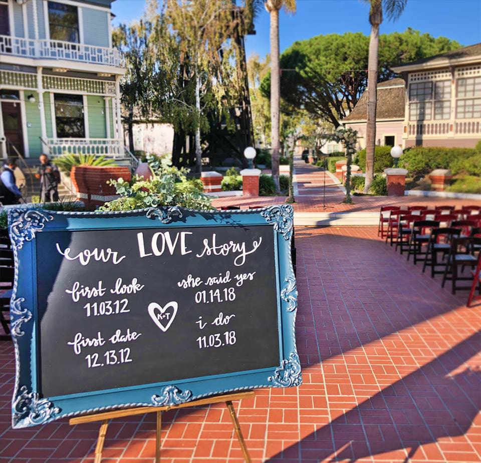 Outdoor wedding venue with chairs and a sign detailing key dates of a couple's relationship, set in a sunny courtyard.