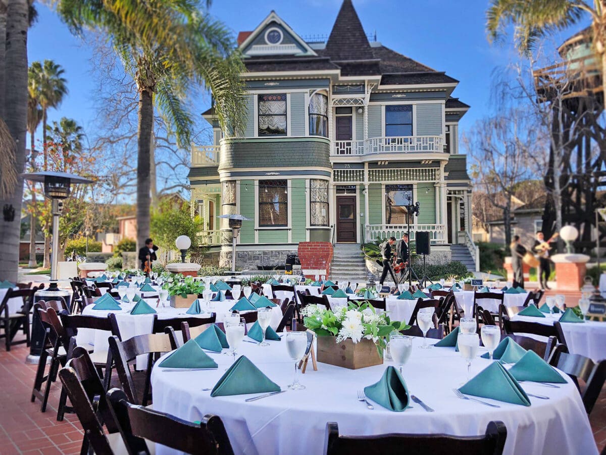 Outdoor event setting with white clothed tables and teal napkins in front of a large, victorian-style green house on a sunny day at Heritage Square, Oxnard.