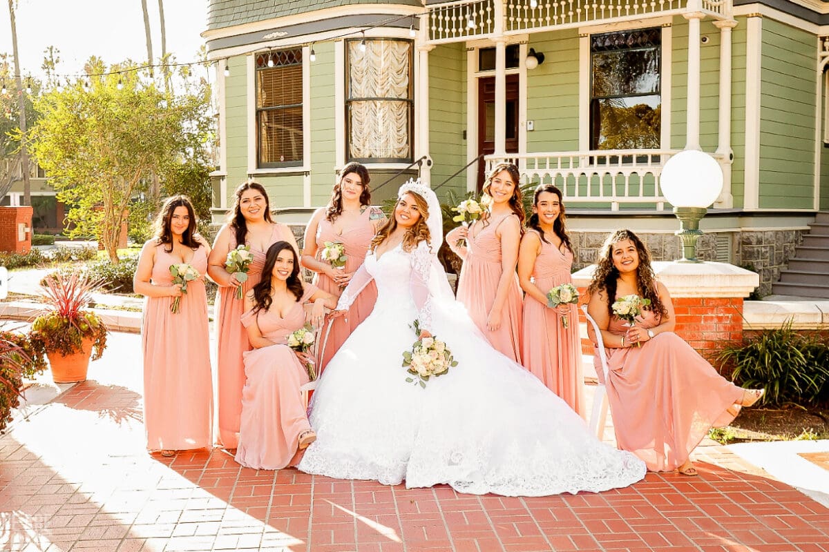 A bride in a white dress stands with seven bridesmaids in matching pink dresses, holding bouquets, outside a charming Victorian house in Heritage Square, Oxnard.