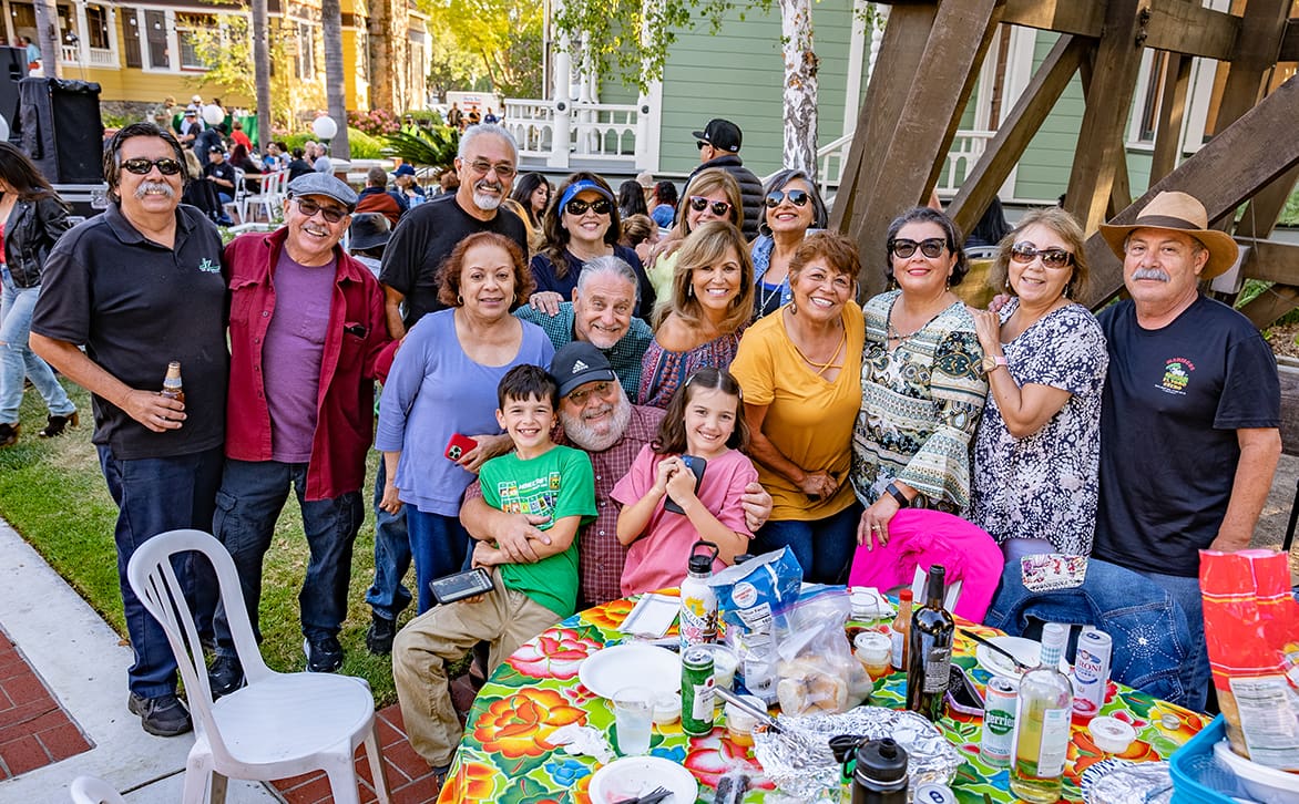 A large, happy multi-generational family gathering at Heritage Square in Oxnard, around a picnic table filled with food, smiling at the camera.