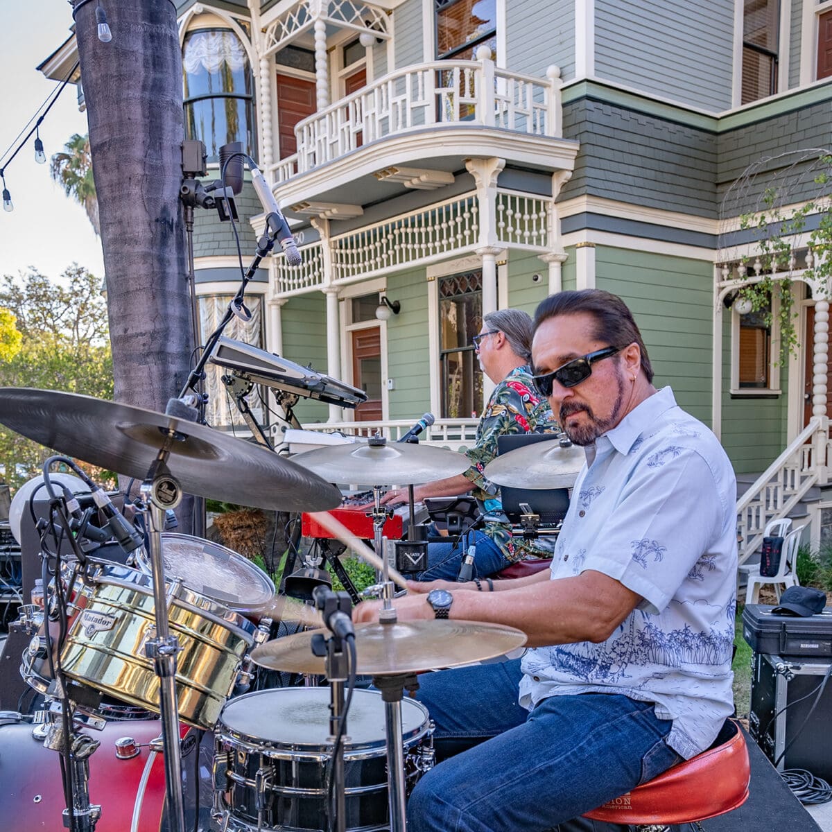 Two male drummers playing kits during an outdoor performance at Heritage Square in front of a Victorian house.