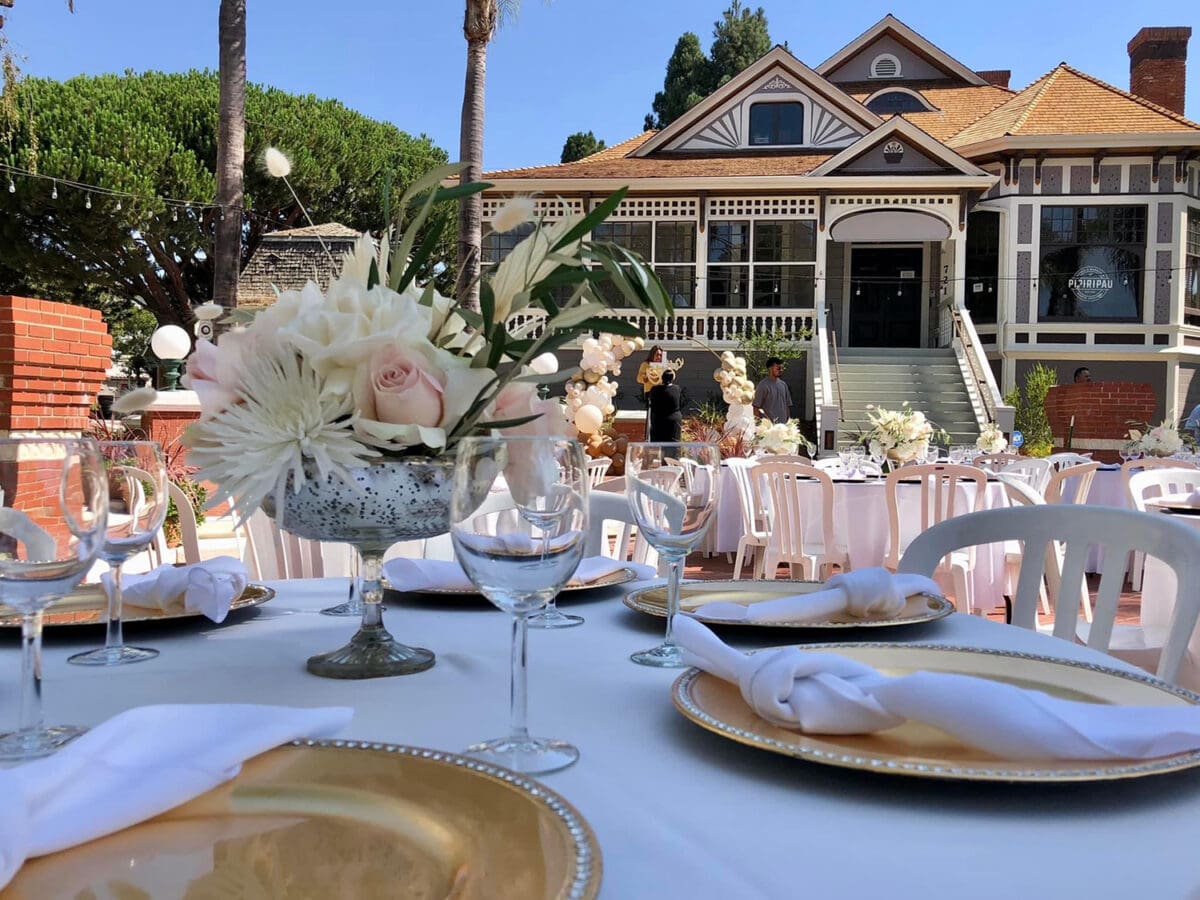 Outdoor wedding reception setup featuring elegantly arranged tables with floral centerpieces in front of a classic Victorian home in Heritage Square, Oxnard on a sunny day.