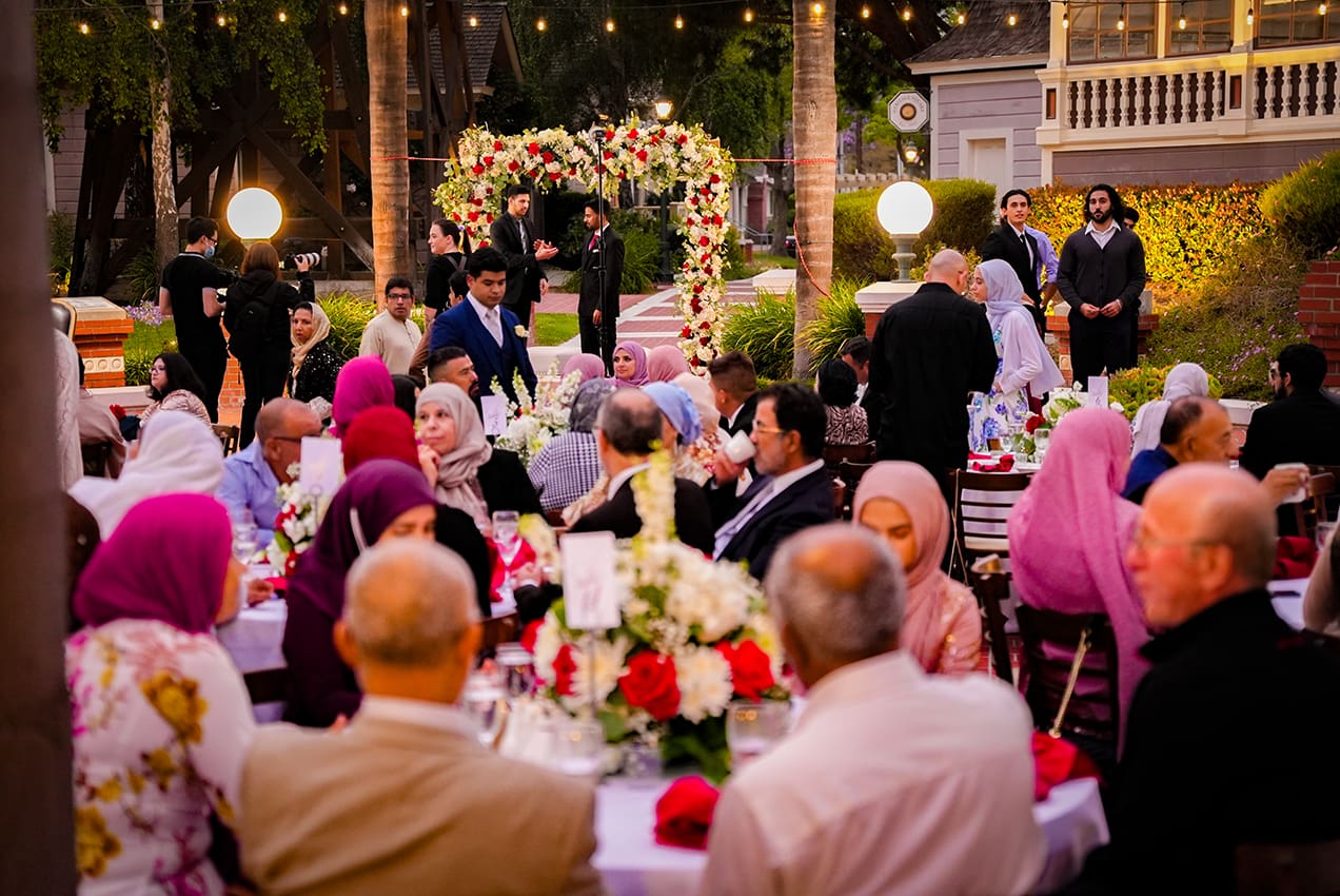 Guests seated at tables during an outdoor wedding reception at Heritage Square, with a floral arch in the background, during evening.