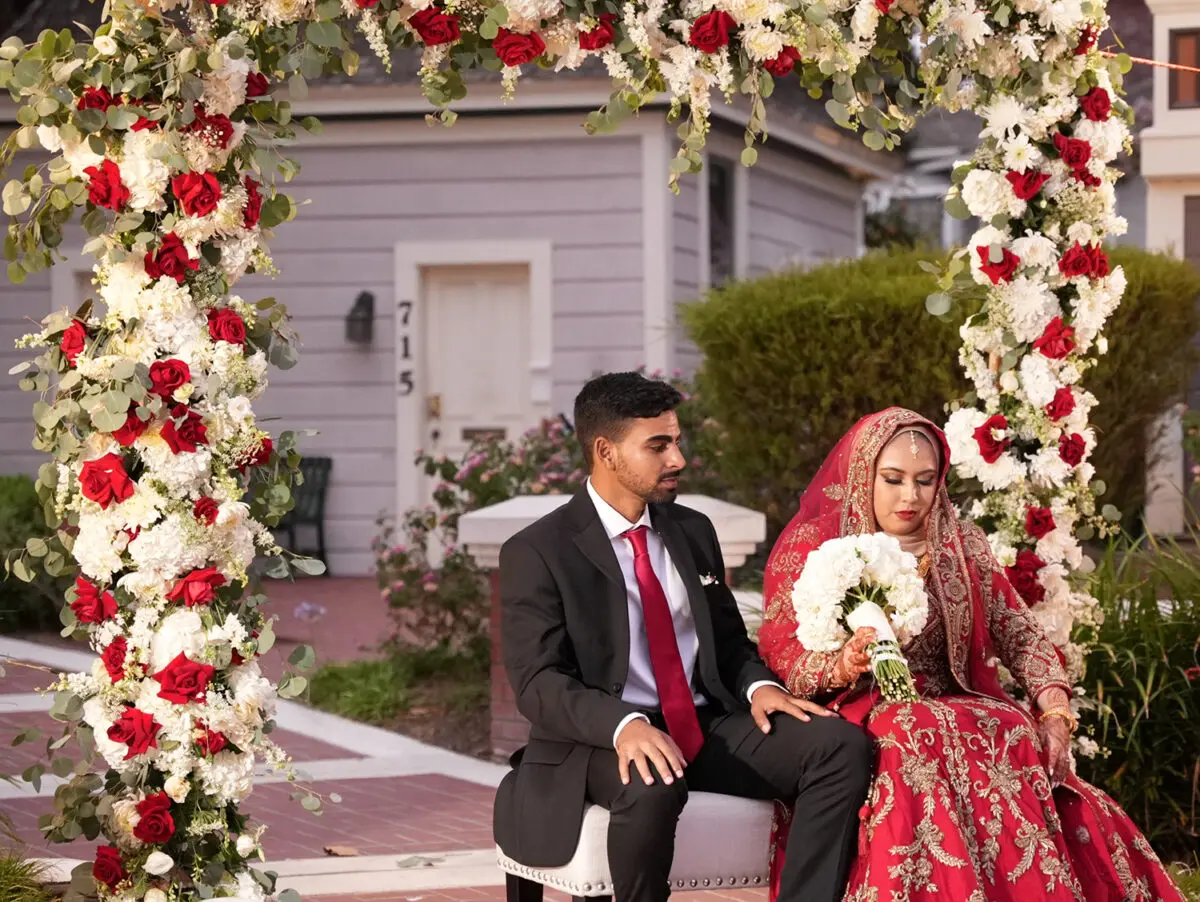 A couple in wedding attire sits under a floral arch at Heritage Square Oxnard, the woman in a red dress and the man in a suit with a red tie.