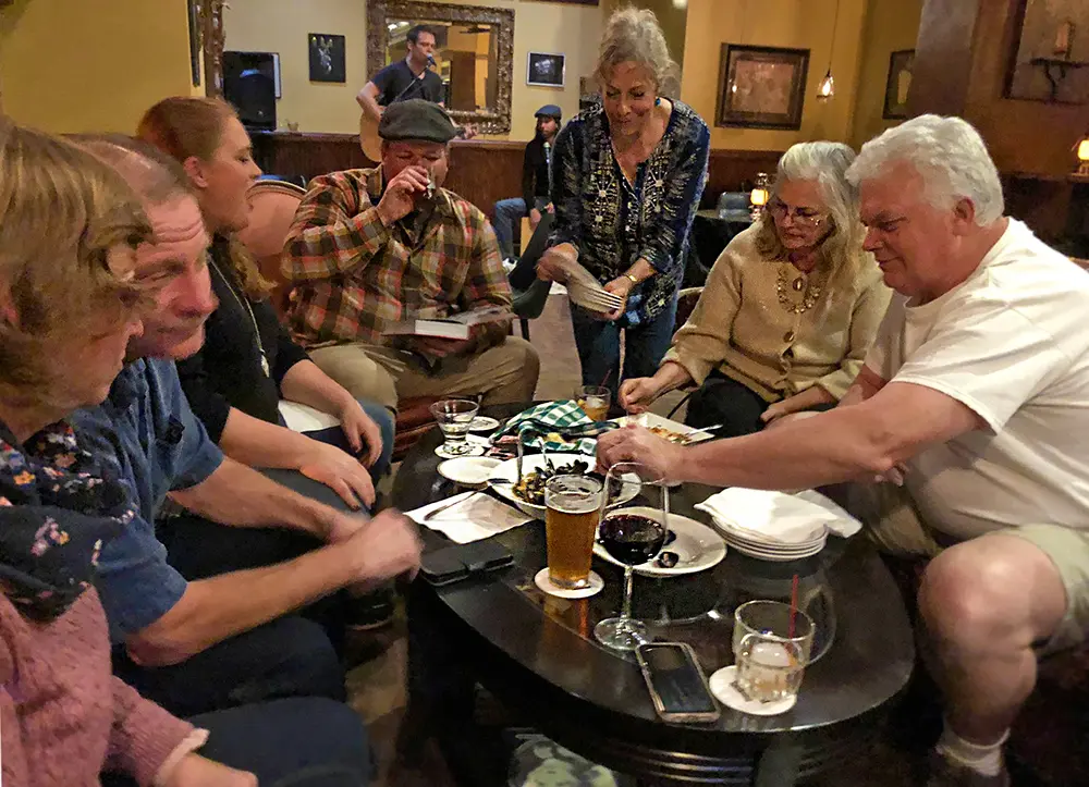 A lively group of older adults enjoying drinks and conversation around a crowded table in a warmly lit pub at Heritage Square.