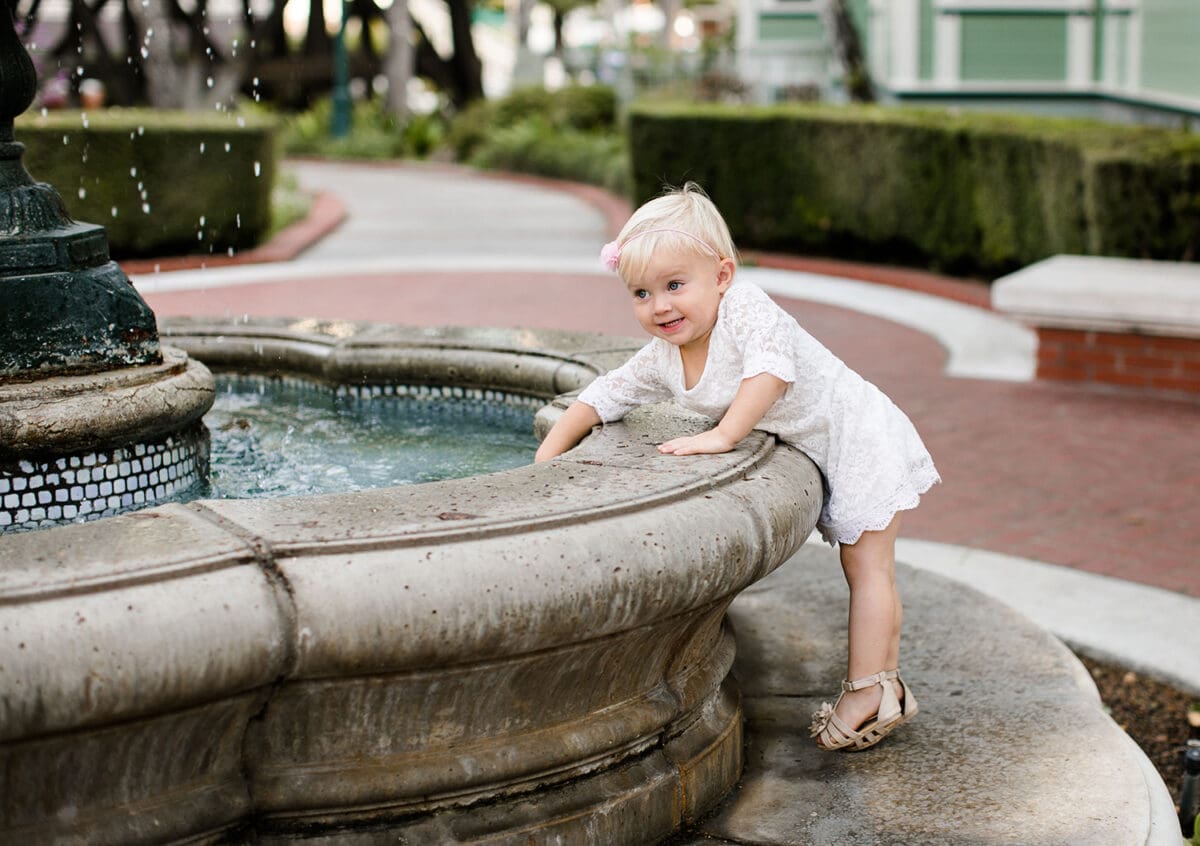 Toddler girl in a white dress leaning over the edge of a fountain at Heritage Square, Oxnard, looking at the water with a playful expression.