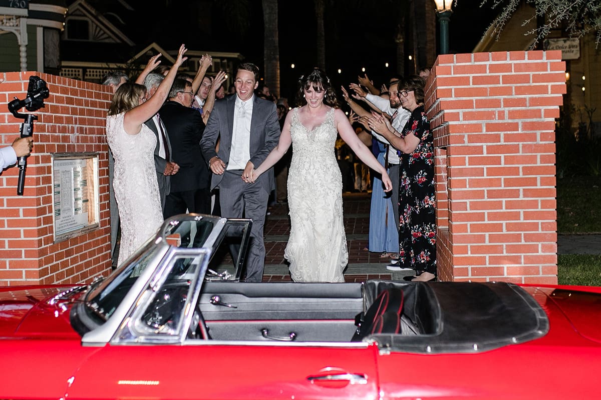 A newly married couple holds hands and walks joyfully through a cheering crowd at Heritage Square toward a red convertible at night.