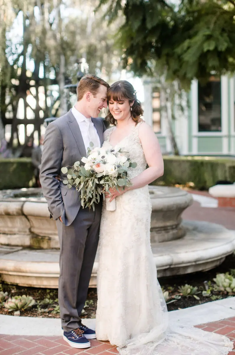A bride and groom smiling at each other, holding a bouquet, standing by a fountain in Heritage Square. The groom is dressed in a gray suit and the bride in a lace gown.