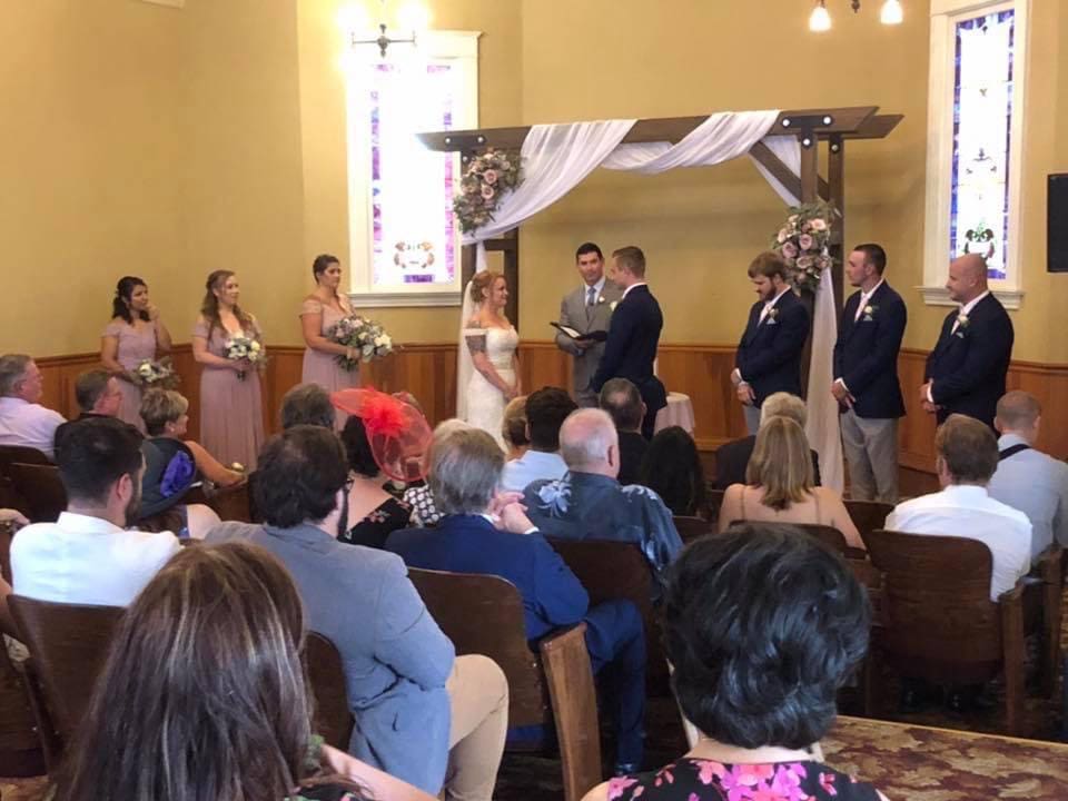 A wedding ceremony in a room with stained glass windows. the bride and groom face each other in front, flanked by bridesmaids and groomsmen, with guests seated watching.