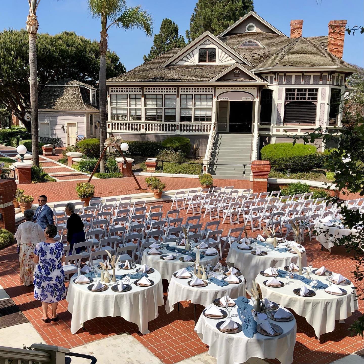 Outdoor wedding setup in Heritage Square in front of a classic Victorian house with rows of white chairs and round tables set for dining under a clear sunny sky.