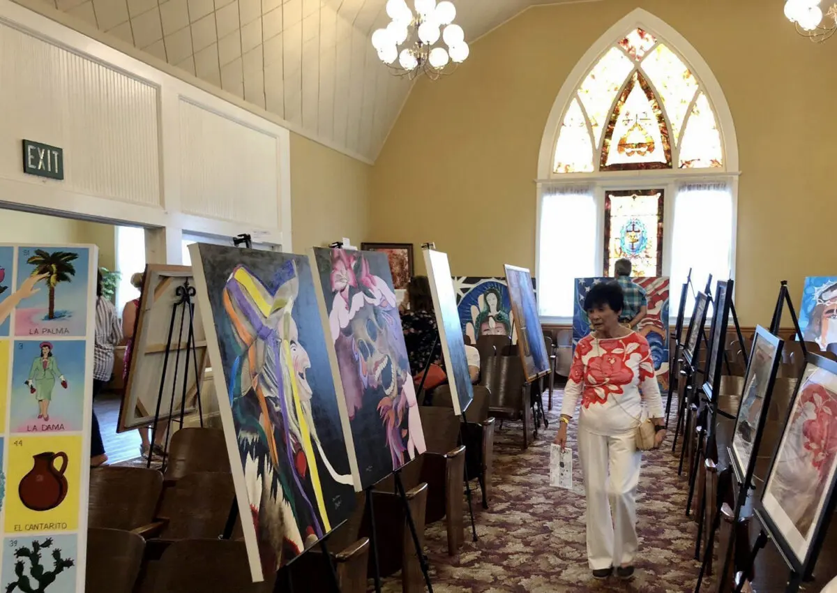 An art exhibit at Heritage Square in Oxnard featuring colorful paintings on easels inside a bright room with arched stained glass windows, and a woman in white clothing observing the artwork.