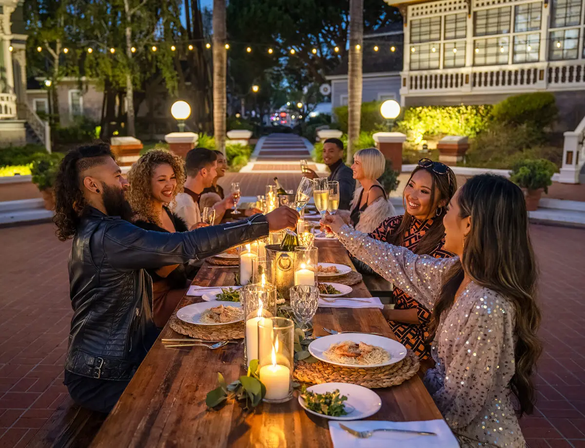 A group of friends enjoying dinner outdoors at Heritage Square, Oxnard, around a candlelit table during the evening, with one man reaching for a wine bottle.