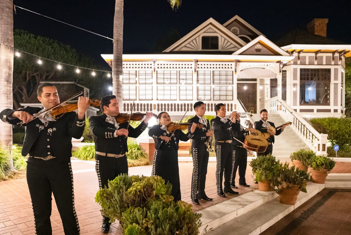 Mariachi band performing at night in Heritage Square with a lit-up exterior and white picket fence.