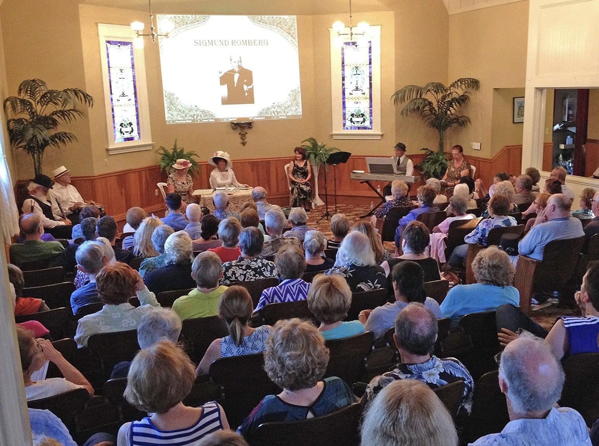 Audience attending a presentation about Siegmund Romberg in Heritage Square, Oxnard, with a speaker in front and a large screen displaying an image of Romberg.
