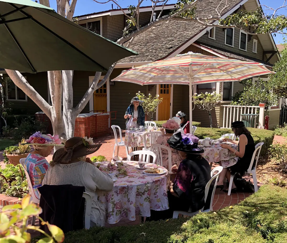 A group of people wearing vintage hats enjoys a tea party at Heritage Square, with tables covered in floral tablecloths, surrounded by lush greenery and a quaint house in the background.