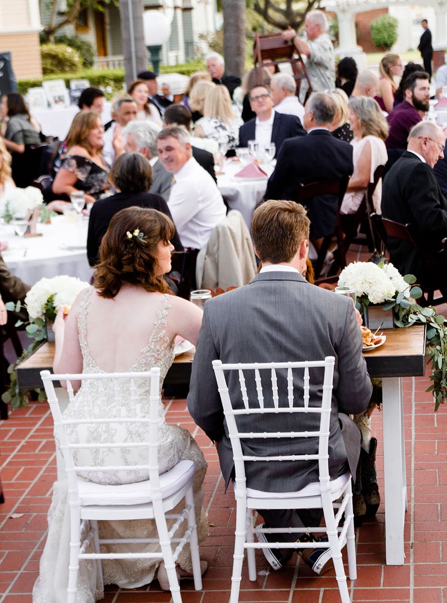 Bride and groom sitting at a reception table in Heritage Square, Oxnard, surrounded by guests, with a violinist playing in the background.