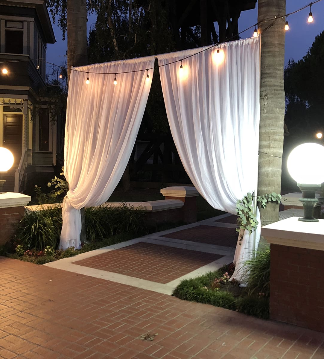 White curtains draped between two palm trees at Heritage Square, Oxnard, lit by soft globe lights, creating an inviting entryway.