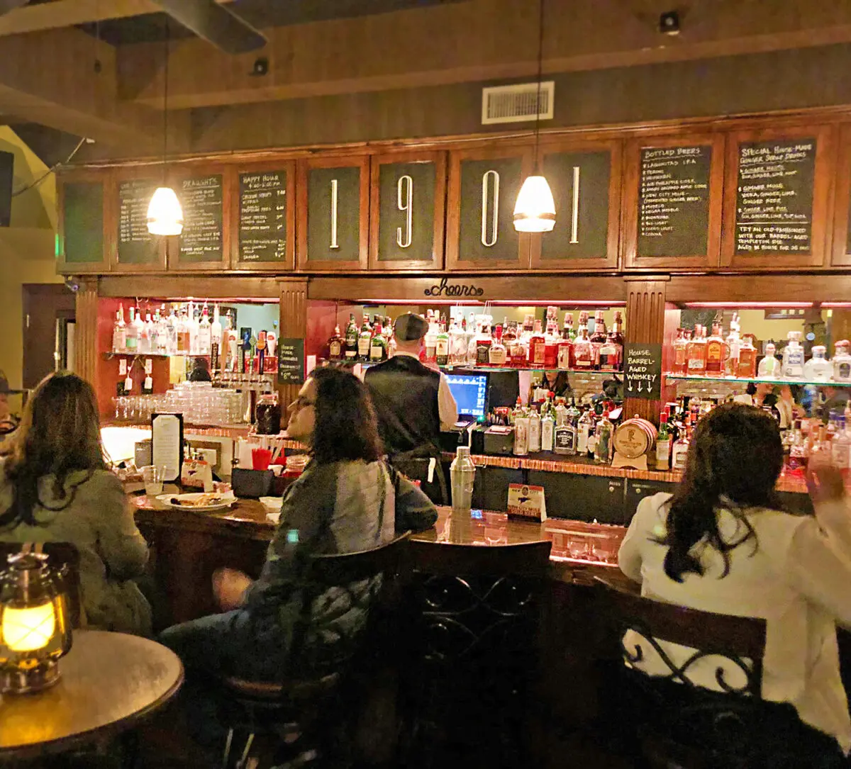 A cozy bar scene in Heritage Square with patrons seated, a bartender at work, and an array of liquor bottles displayed under a sign marked "1904".
