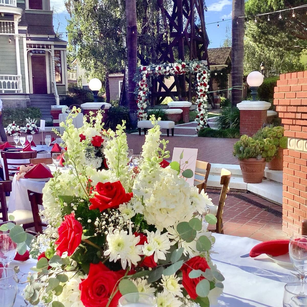 Outdoor wedding reception setup with floral centerpieces, red napkins, and an arch decorated with flowers in a garden courtyard.