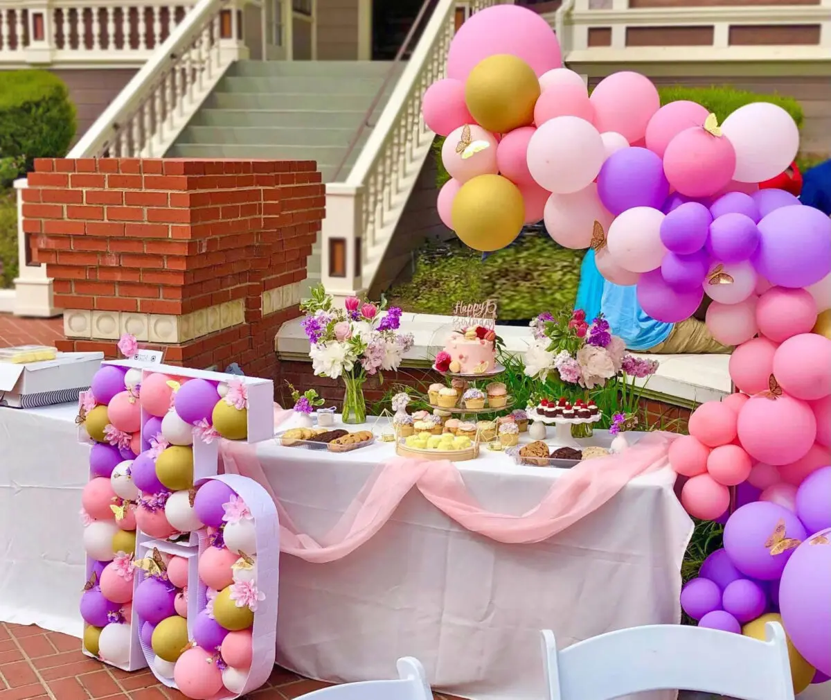 Outdoor birthday party setup at Heritage Square, Oxnard, with a table of desserts, colorful balloons, and white chairs in front of a house.