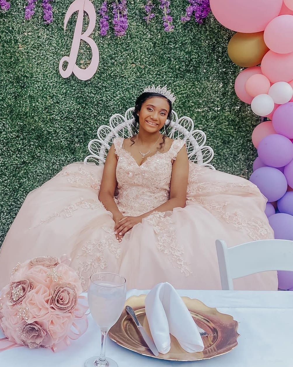 A woman in a pink gown and tiara sits at a table decorated with a balloon arch and flowers at Heritage Square in Oxnard, smiling at a celebration event.