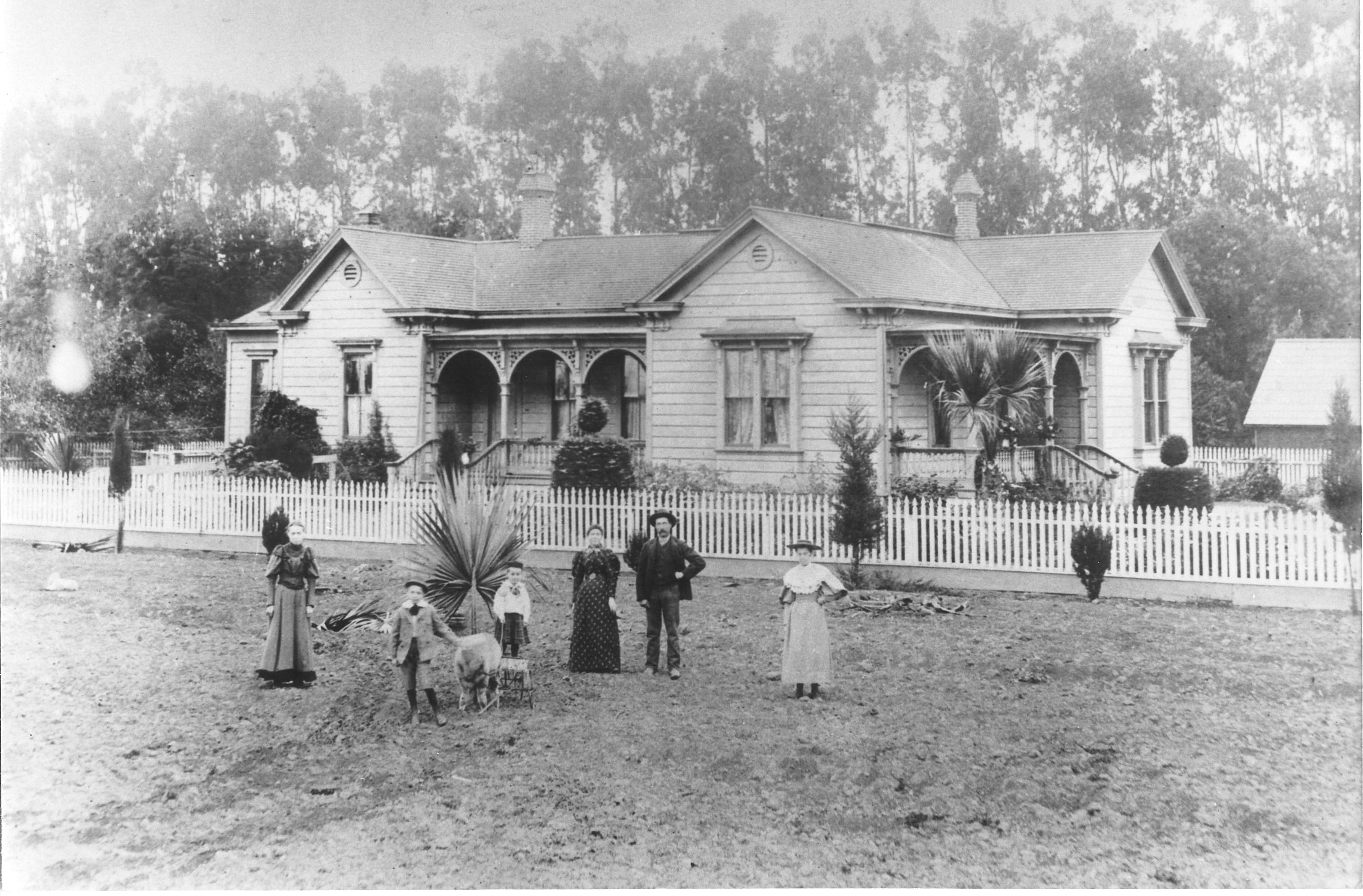 Historic black-and-white photo of a family and two dogs in front of a 19th-century house in Heritage Square, Oxnard, with a picket fence and lush garden.
