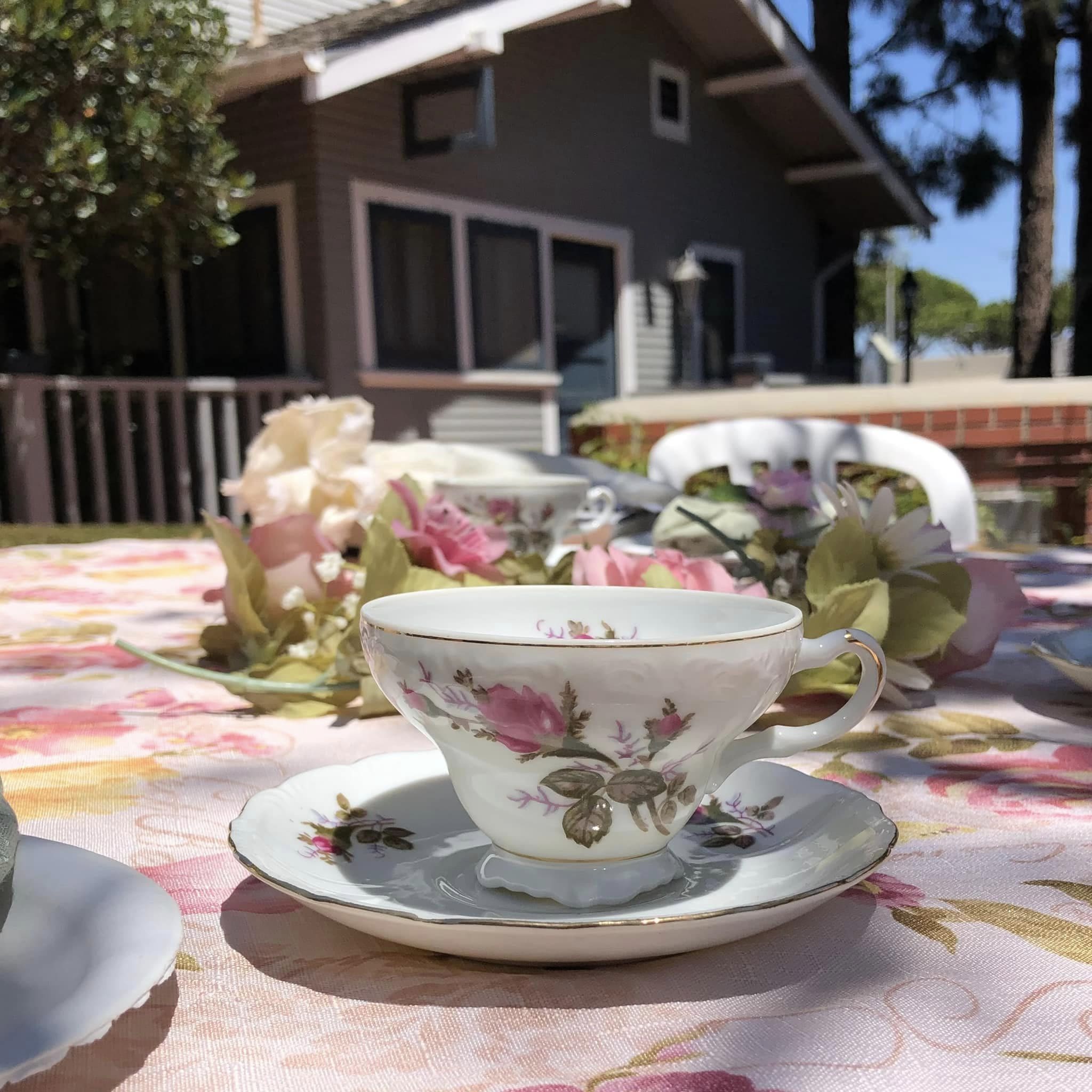 A porcelain teacup with floral design on a saucer, set on a table with a flower-patterned tablecloth, in an outdoor setting at Heritage Square with a house and trees in the background