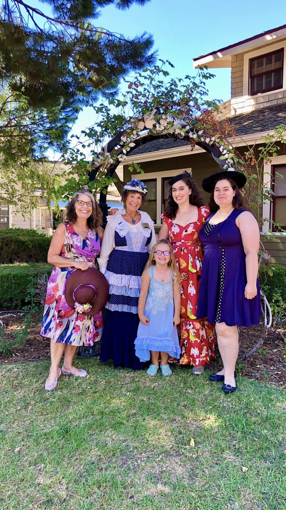 Five women in colorful dresses and one young girl posing in front of a quaint garden arch at Heritage Square, Oxnard, on a sunny day.