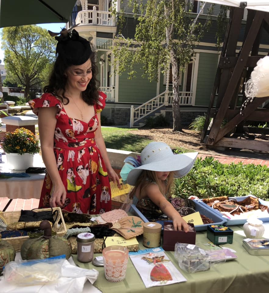 A woman in a red floral dress and a hat smiles while standing at a table with various items at Heritage Square, Oxnard, as a young girl in a wide-brimmed hat examines the