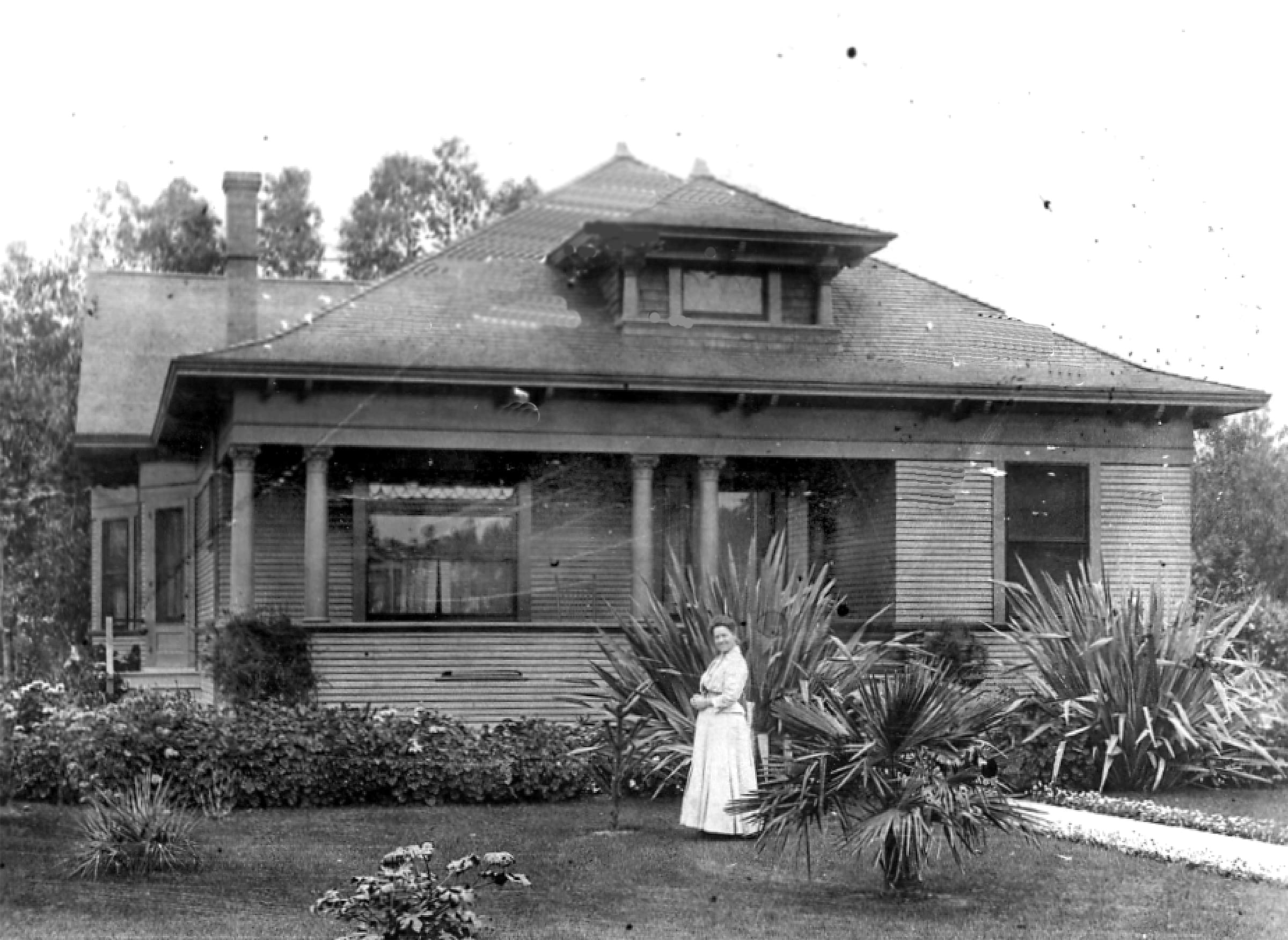A vintage black-and-white photo of a person in a white dress standing in front of a classic house at Heritage Square, Oxnard, with a lush garden and tall palm plants.
