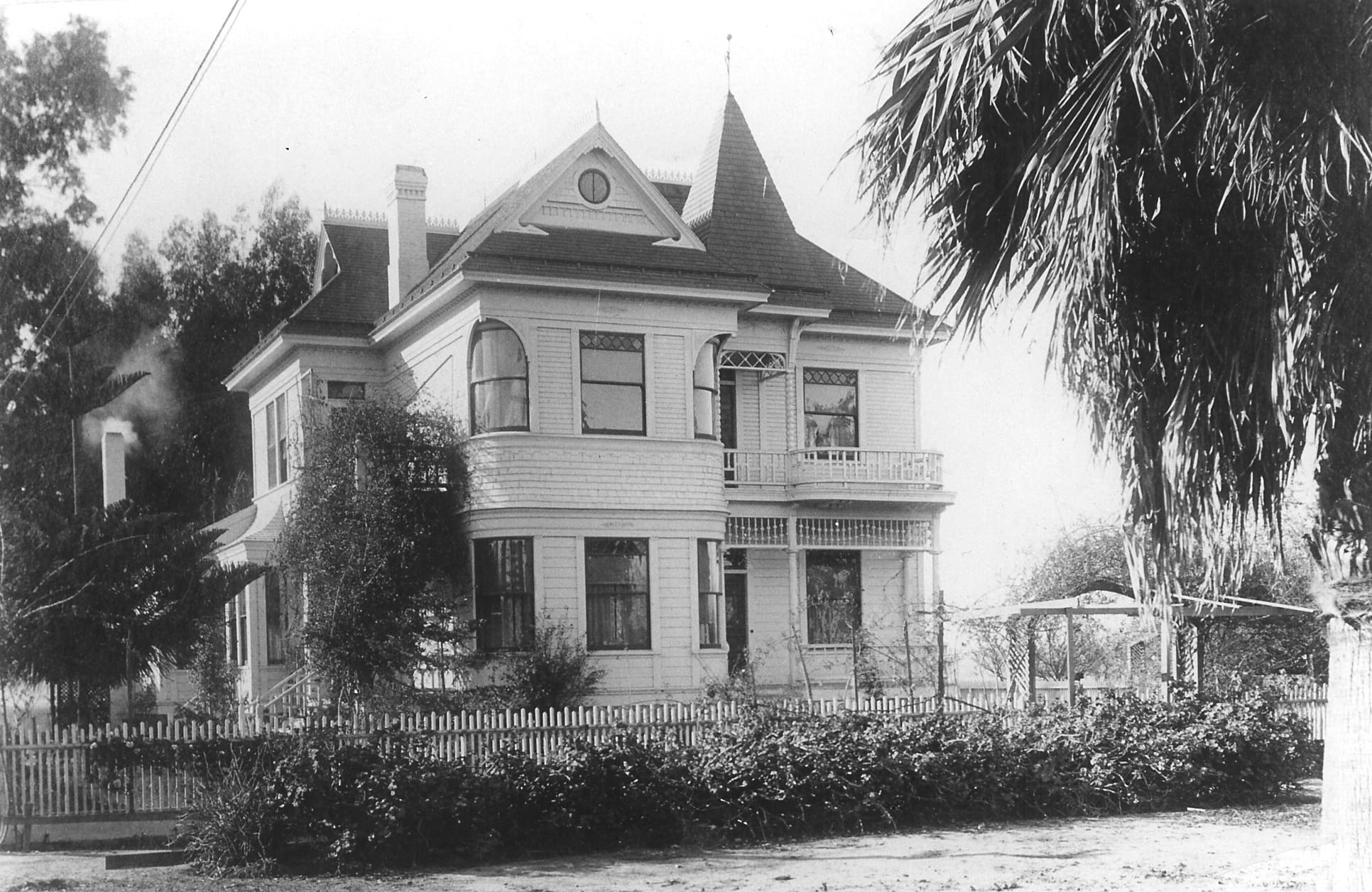 Black and white historical photo of a two-story Victorian house with a turret in Oxnard, surrounded by a picket fence and palm trees.