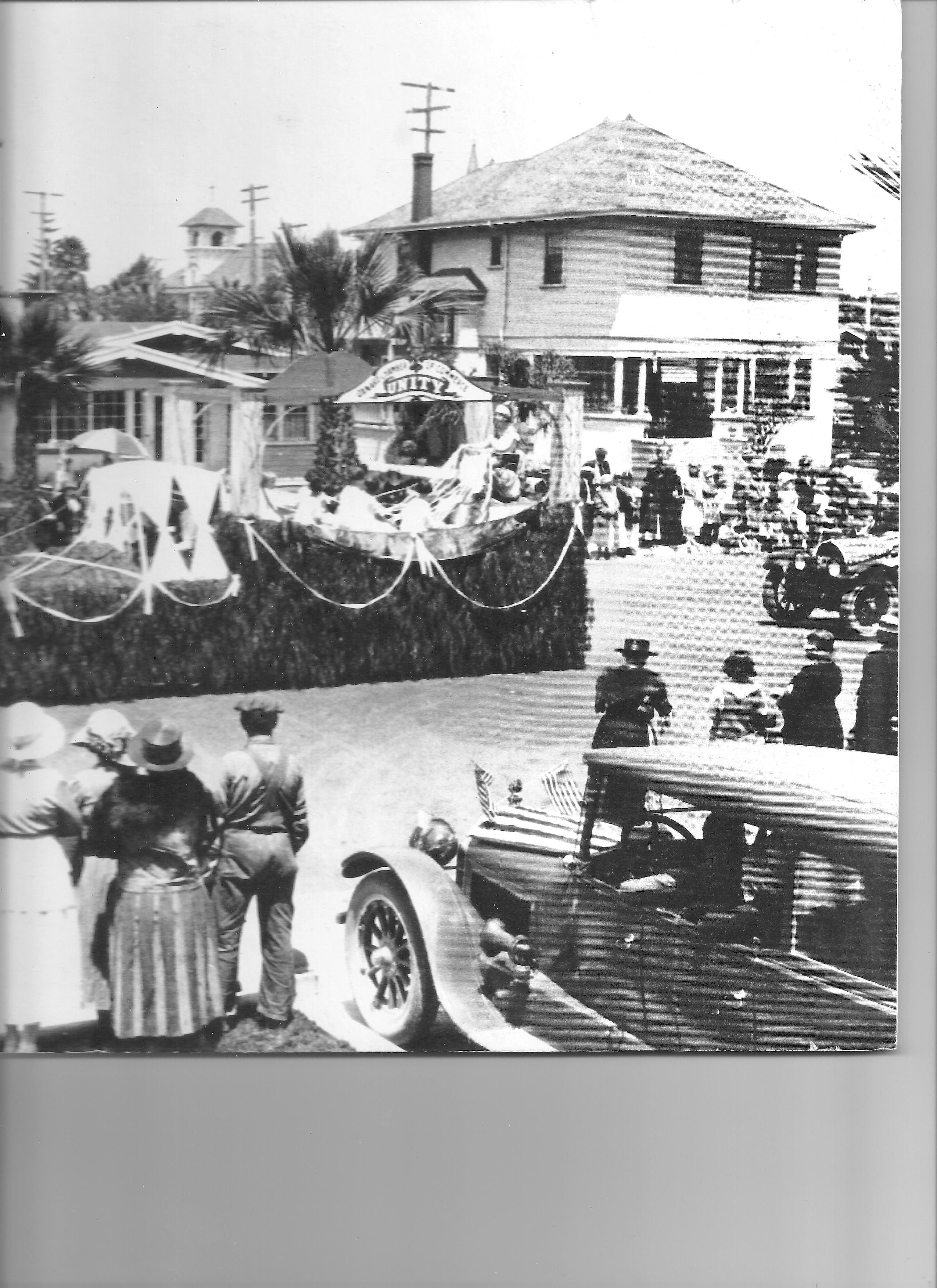 Historical black and white photograph of a parade with spectators and vintage cars, featuring a decorated float and nearby buildings.