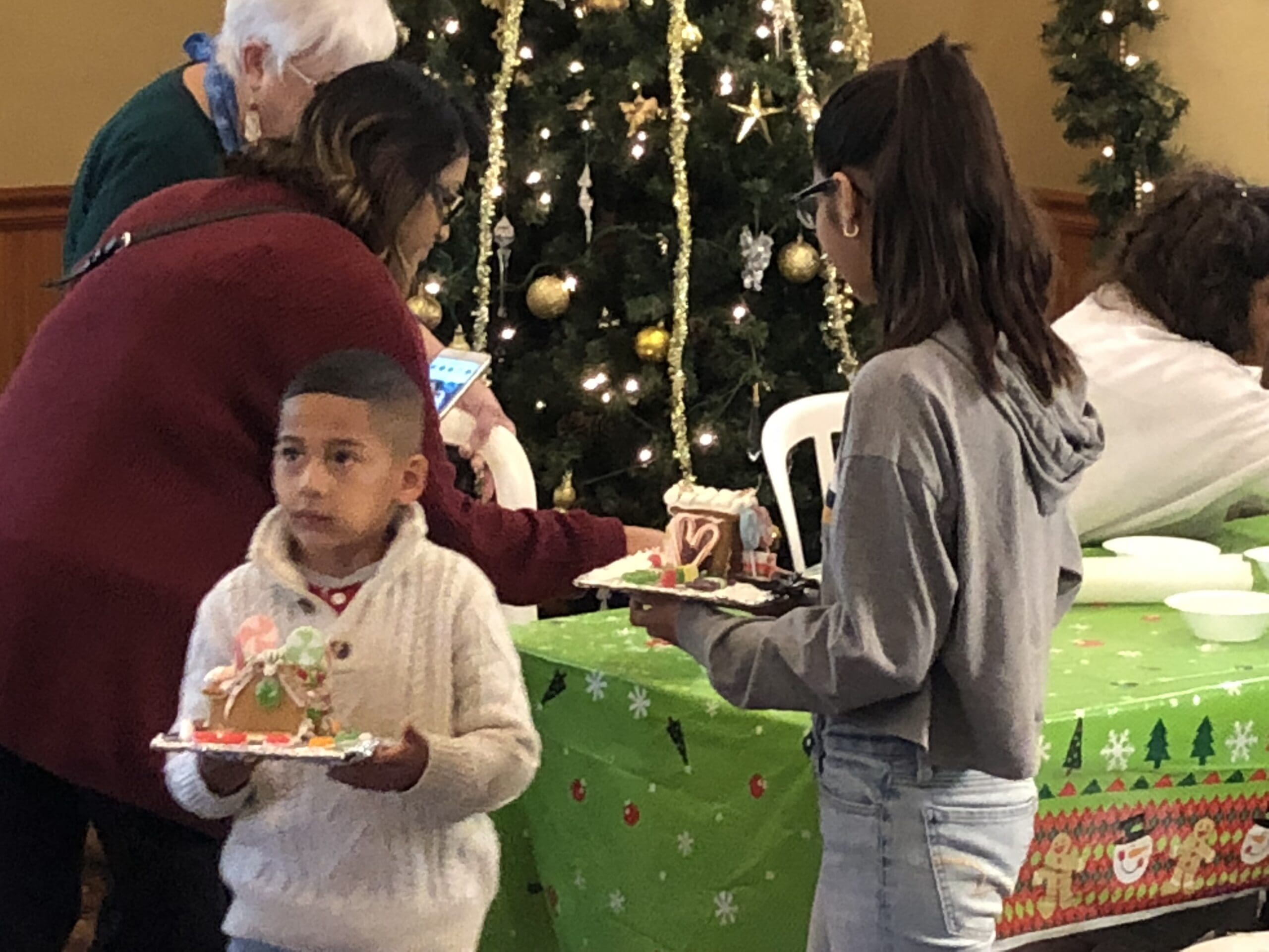Two children holding gingerbread houses at a Christmas party in Heritage Square, with a decorated tree in the background and other people around.