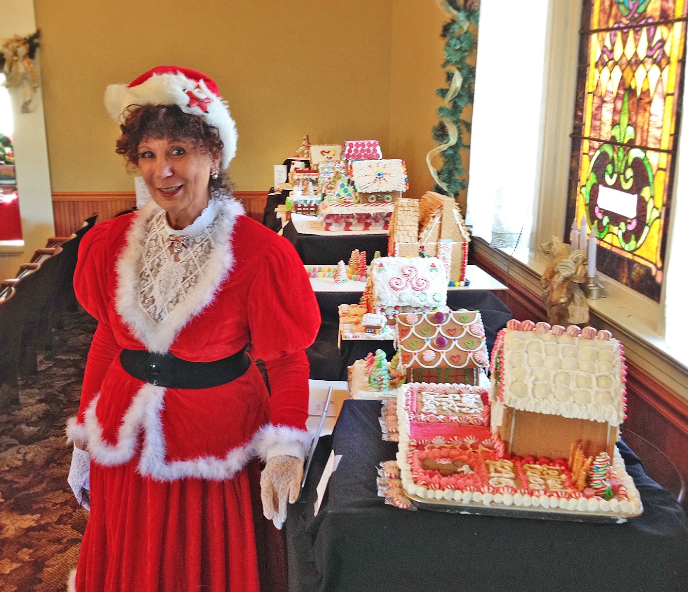 Woman dressed as Mrs. Claus standing next to a table of gingerbread houses in a festively decorated room at Heritage Square, Oxnard.