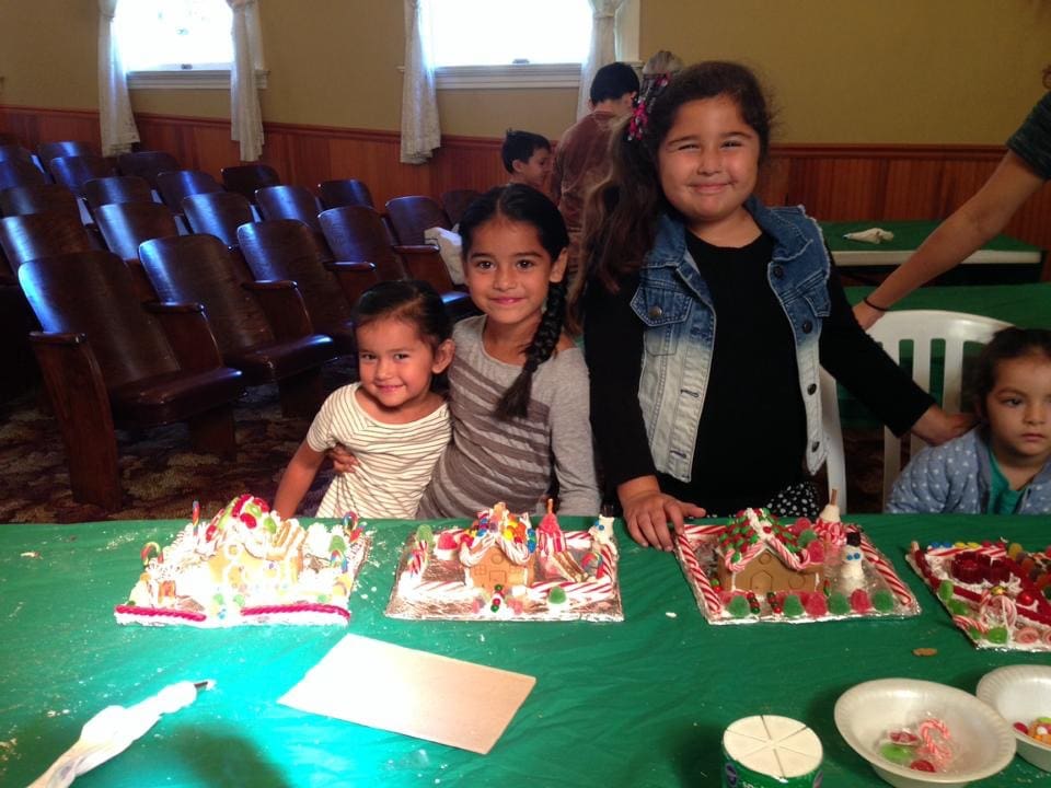 Three children smiling beside their gingerbread houses on a table at a crafting event in Heritage Square, Oxnard.