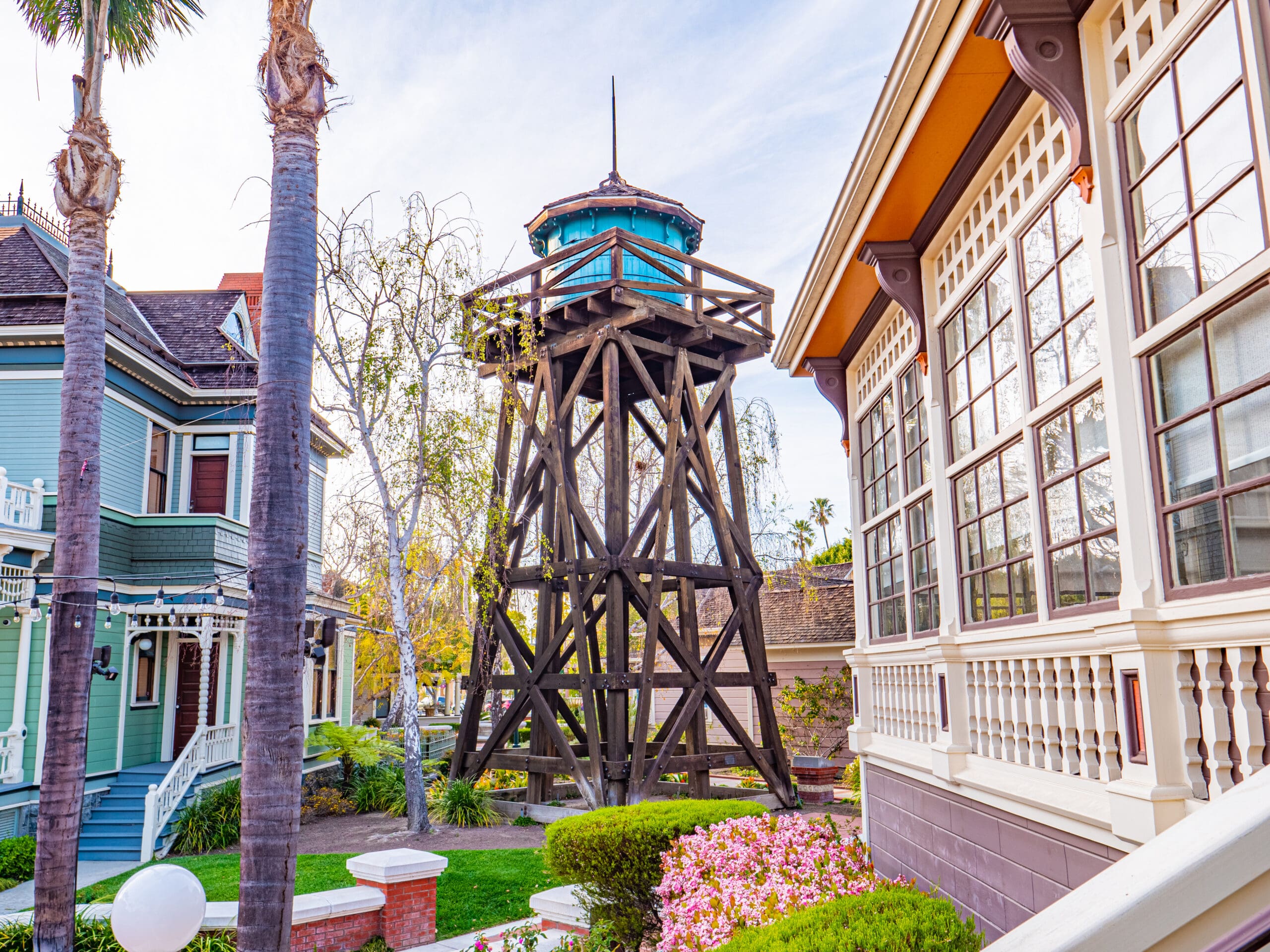 Wooden water tower between two Victorian-style houses with manicured garden and palm trees in a sunny Oxnard suburb setting.