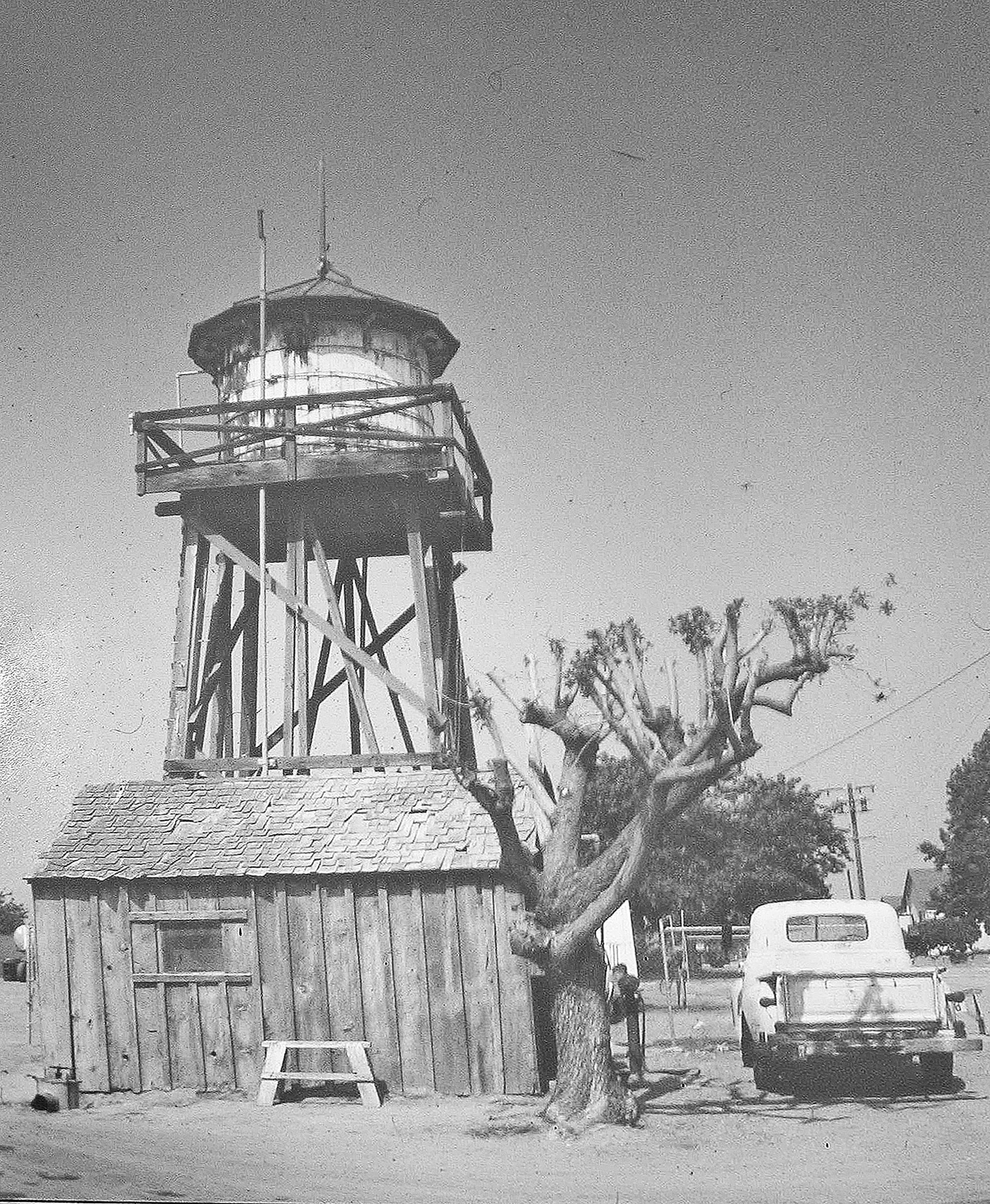 Black and white photo of an old wooden water tower above a small shack at Heritage Square, with a leafless tree and an old van parked nearby.