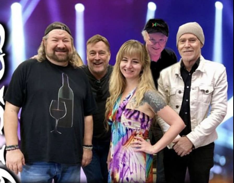 A group of five people standing together and smiling in front of a colorful, illuminated backdrop. One person has long hair and a beard, and another person wears a hat.