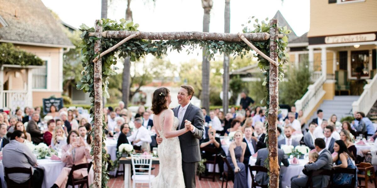 A bride and groom share a dance under a rustic arch, surrounded by seated guests at an outdoor wedding venue in Heritage Square, Oxnard.